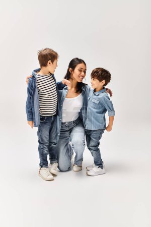 Eine junge asiatische Mutter und ihre beiden kleinen Söhne stehen zusammen in einem grauen Studio, alle in Jeans gekleidet.