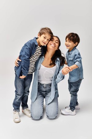Téléchargez les photos : Une jeune mère asiatique et ses petits fils, tous vêtus de denim, posant ensemble pour un portrait dans un studio gris. - en image libre de droit