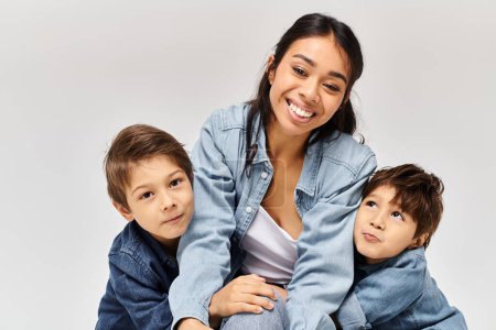 Eine junge asiatische Mutter sitzt auf ihren beiden kleinen Söhnen, alle in Jeanskleidung in einem grauen Studio-Setting.