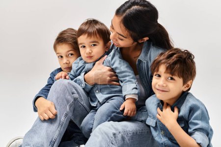 Eine junge asiatische Mutter in Jeanskleidung sitzt auf ihren kleinen Söhnen, ebenfalls in Jeans, alle in einem grauen Studio.