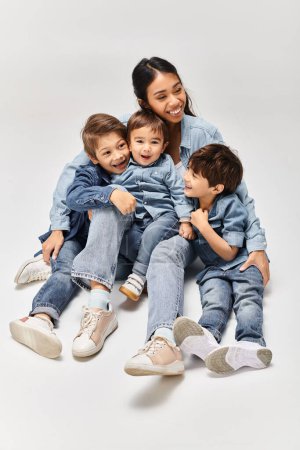 Eine junge asiatische Mutter sitzt majestätisch auf einer Gruppe von Kindern, alle in Jeanskleidung, in einer grauen Studiokulisse.