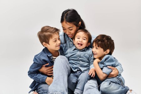 A group of children, including a young Asian mother and her sons, in denim clothes, sit on top of each other in a grey studio.