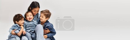Téléchargez les photos : A group of children, including young Asian mother and her sons, playfully stack atop one another in a grey studio setting. - en image libre de droit