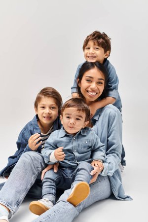 Foto de Una joven madre asiática y sus hijos pequeños se apilan juguetonamente uno encima del otro, todos vistiendo ropa de mezclilla en un estudio gris. - Imagen libre de derechos