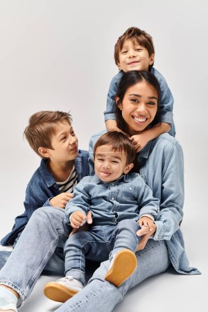 Foto de Un grupo de personas, una joven madre asiática y sus hijos pequeños, sentados uno encima del otro en un estudio gris, todos con ropa de mezclilla. - Imagen libre de derechos