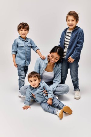 Foto de Una joven madre asiática y sus hijos pequeños, todos vestidos con denim, posan juntos en un estudio gris. - Imagen libre de derechos