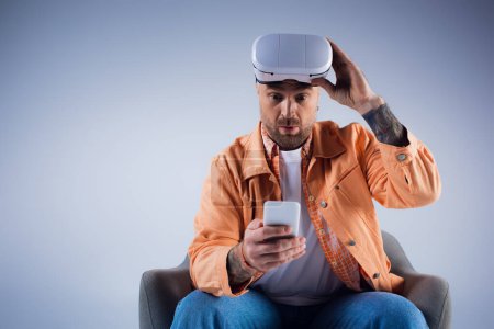 Téléchargez les photos : Un homme sur une chaise tient un smartphone dans un décor studio, immergé dans le monde de la métaverse à travers son casque VR. - en image libre de droit