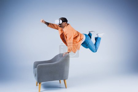 Foto de Un hombre con una camisa naranja mostrando un truco que desafía la gravedad mientras se equilibra en una silla en un entorno de estudio. - Imagen libre de derechos