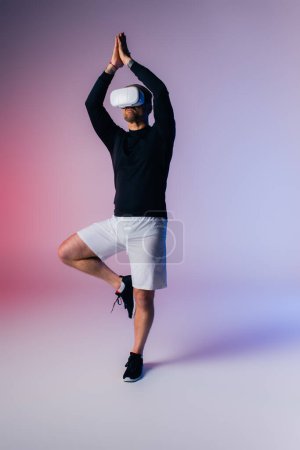Un hombre con una camisa negra y pantalones cortos blancos practica una postura desafiante de yoga con equilibrio y control en un entorno de estudio.