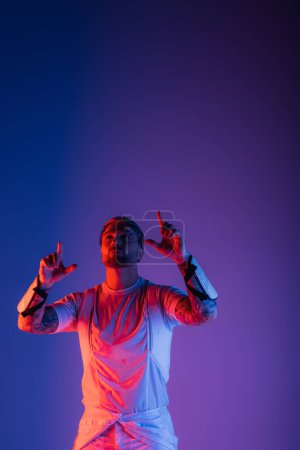 Foto de Un hombre con gafas inteligentes se levanta con confianza contra un vibrante telón de fondo púrpura y azul en un entorno de estudio. - Imagen libre de derechos
