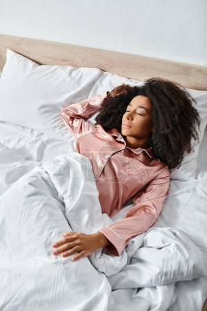 Eine lockige Afroamerikanerin im Schlafanzug legt sich morgens friedlich auf ein weißes Bett in einem sonnenbeschienenen Schlafzimmer.