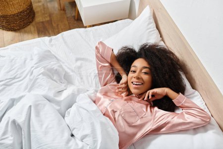 Curly African American woman in pajamas peacefully resting on a white bed in a serene bedroom during the morning.