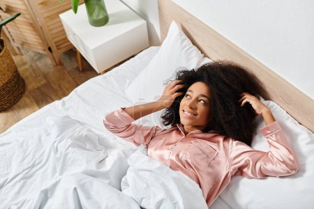 Una mujer afroamericana rizada en pijama descansando pacíficamente sobre una cama blanca por la mañana.