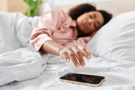 Foto de Mujer afroamericana rizada en pijama relajándose en la cama, absorta en su teléfono celular por la mañana. - Imagen libre de derechos