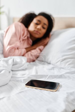 Lockige Afroamerikanerin im Pyjama, friedlich im Bett liegend neben einem Handy in einem gemütlichen Schlafzimmer während des Morgens.