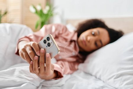 Curly African American woman in pajamas laying in bed, holding a remote control in her hand, relaxing in the morning.
