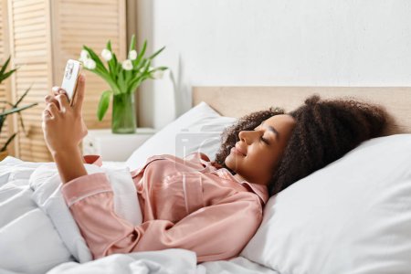 A curly African American woman in pajamas lays in bed, holding a cell phone