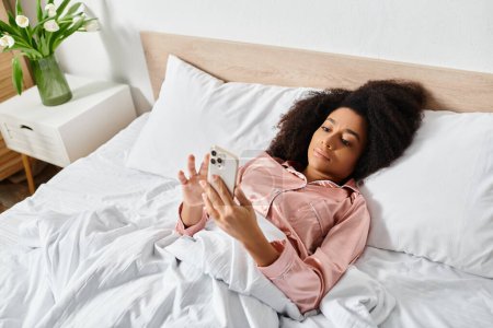 Photo for Curly African American woman in pajamas relaxes on bed, holding cell phone in morning light. - Royalty Free Image