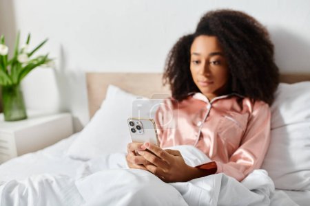 Curly African American woman in pajamas laying in bed, engrossed with cell phone in early morning light.