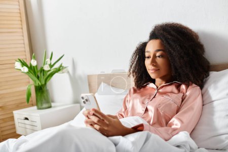 Femme afro-américaine bouclée en pyjama assise sur le lit, absorbée dans son téléphone, lumière du matin filtrant par la fenêtre.