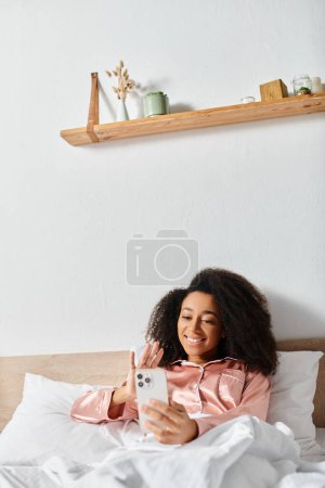 Photo for A curly African American woman in pajamas takes a selfie with her phone while laying in bed in the morning. - Royalty Free Image