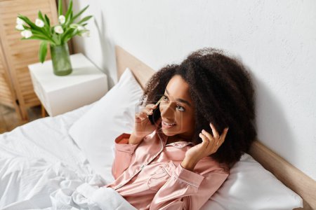 Foto de Mujer afroamericana rizada en pijama hablando por teléfono celular mientras está acostada en la cama en la mañana. - Imagen libre de derechos