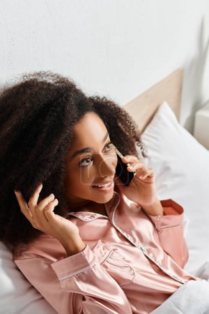 A curly African American woman in pajamas talking on a cell phone while laying in bed in the morning.