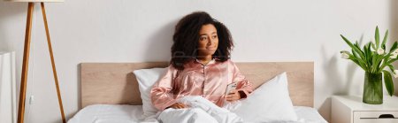A curly African American woman in pajamas sits peacefully on a bed next to fluffy pillows in a cozy bedroom during morning time.