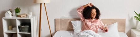 Foto de Una mujer afroamericana rizada se relaja en la parte superior de una cama en su dormitorio, vistiendo una camisa rosa durante la tranquila mañana. - Imagen libre de derechos