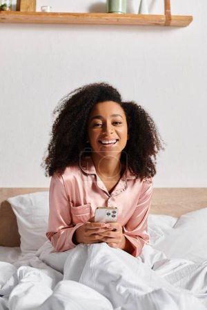 Curly African American woman in pajamas sitting on bed, engrossed in her cell phone screen in the morning.