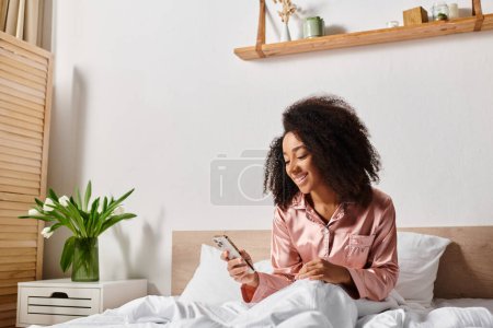 Curly African American woman in pajamas sits on bed, deep in thought, focusing on her cell phone screen in the morning.