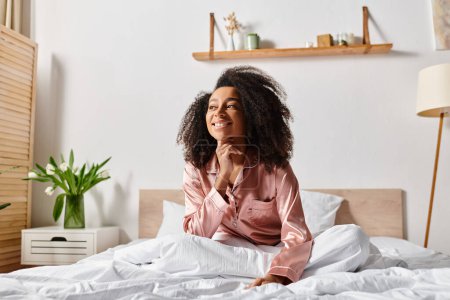 A peaceful scene of a curly African American woman in pajamas sitting on a bed with white sheets in a bright morning bedroom.