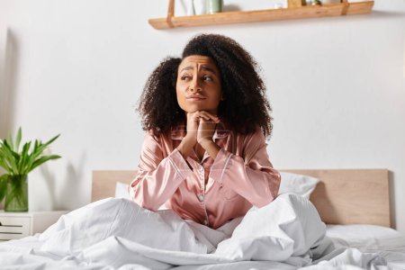 Téléchargez les photos : Une afro-américaine frisée en pyjama s'assoit paisiblement sur un lit blanc dans un cadre matinal serein. - en image libre de droit