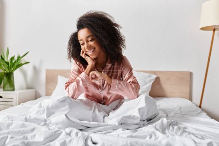 A curly African American woman in pajamas sits calmly on a bed with white sheets in a serene bedroom in the morning.