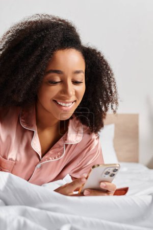 Lockige Afroamerikanerin im Schlafanzug im Bett liegend, morgens von ihrem Handy-Bildschirm gefesselt.