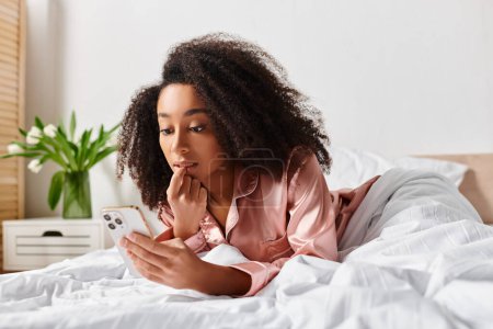 Eine lockige Afroamerikanerin im Schlafanzug liegt im Bett, vertieft, als sie an einem faulen Morgen auf ihr Handy blickt..