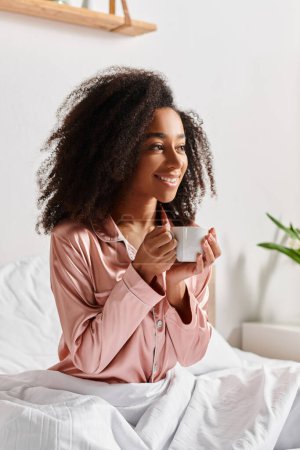 Curly African American woman in pajamas sits on a bed, holding a cup of coffee in a cozy bedroom during morning time.