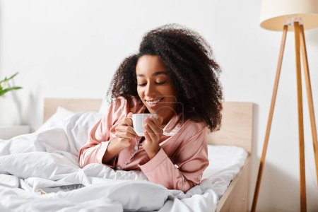 Foto de Una mujer afroamericana rizada en pijama está acostada en la cama, saboreando una taza de café durante la mañana.. - Imagen libre de derechos