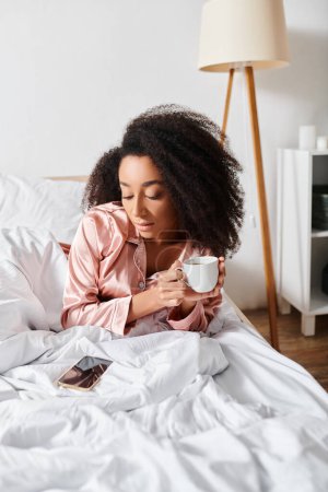 Mujer afroamericana rizada en pijama disfruta de una mañana tranquila con una taza de café en su acogedor dormitorio.