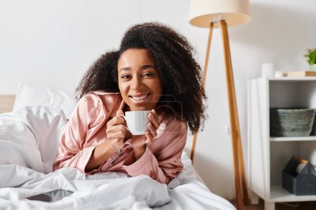 Lockige Afroamerikanerin im Schlafanzug, eingebettet im Bett, ihren Morgen mit einer Tasse Kaffee genießend.