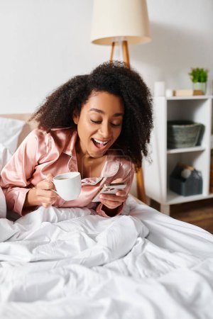 Mujer afroamericana rizada en pijama relajándose en la cama, saboreando una taza de café durante las horas de la mañana.