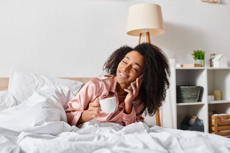 Foto de Una mujer afroamericana con el pelo rizado en pijama, saboreando una taza de café en un acogedor dormitorio durante la mañana. - Imagen libre de derechos