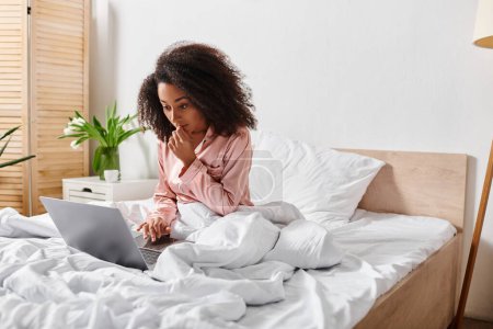 Photo for Curly African American woman in pajamas busy on laptop while sitting on bed in cozy morning setting. - Royalty Free Image
