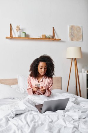Curly African American woman in pajamas sitting on bed, engrossed in laptop screen in a cozy bedroom setting.