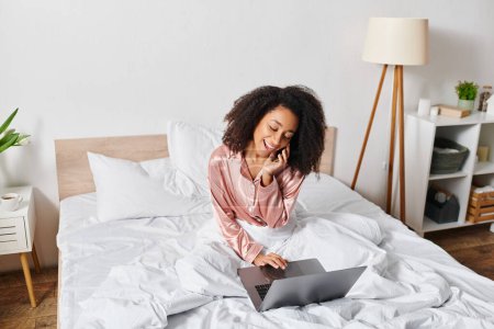Téléchargez les photos : Femme afro-américaine bouclée en pyjama assis sur le lit, absorbé dans l'écran d'ordinateur portable, dans un cadre confortable de chambre à coucher pendant l'heure du matin. - en image libre de droit