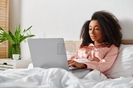 A curly African American woman in pajamas sits on a bed, absorbed in working on a laptop in the morning.