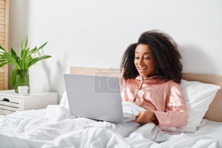Téléchargez les photos : Femme afro-américaine en pyjama assis sur le lit, en utilisant un ordinateur portable dans la chambre le matin. - en image libre de droit