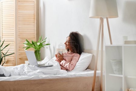Femme afro-américaine bouclée en pyjama assise sur le lit, absorbée dans l'écran d'ordinateur portable, lumière du matin filtrant par la fenêtre.
