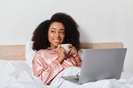 Téléchargez les photos : Femme afro-américaine bouclée en pyjama profitant du matin avec ordinateur portable et café au lit. - en image libre de droit
