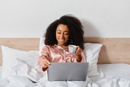 Photo for Curly African American woman in pajamas sitting on bed, holding a cup of coffee, looking at laptop screen in bedroom. - Royalty Free Image